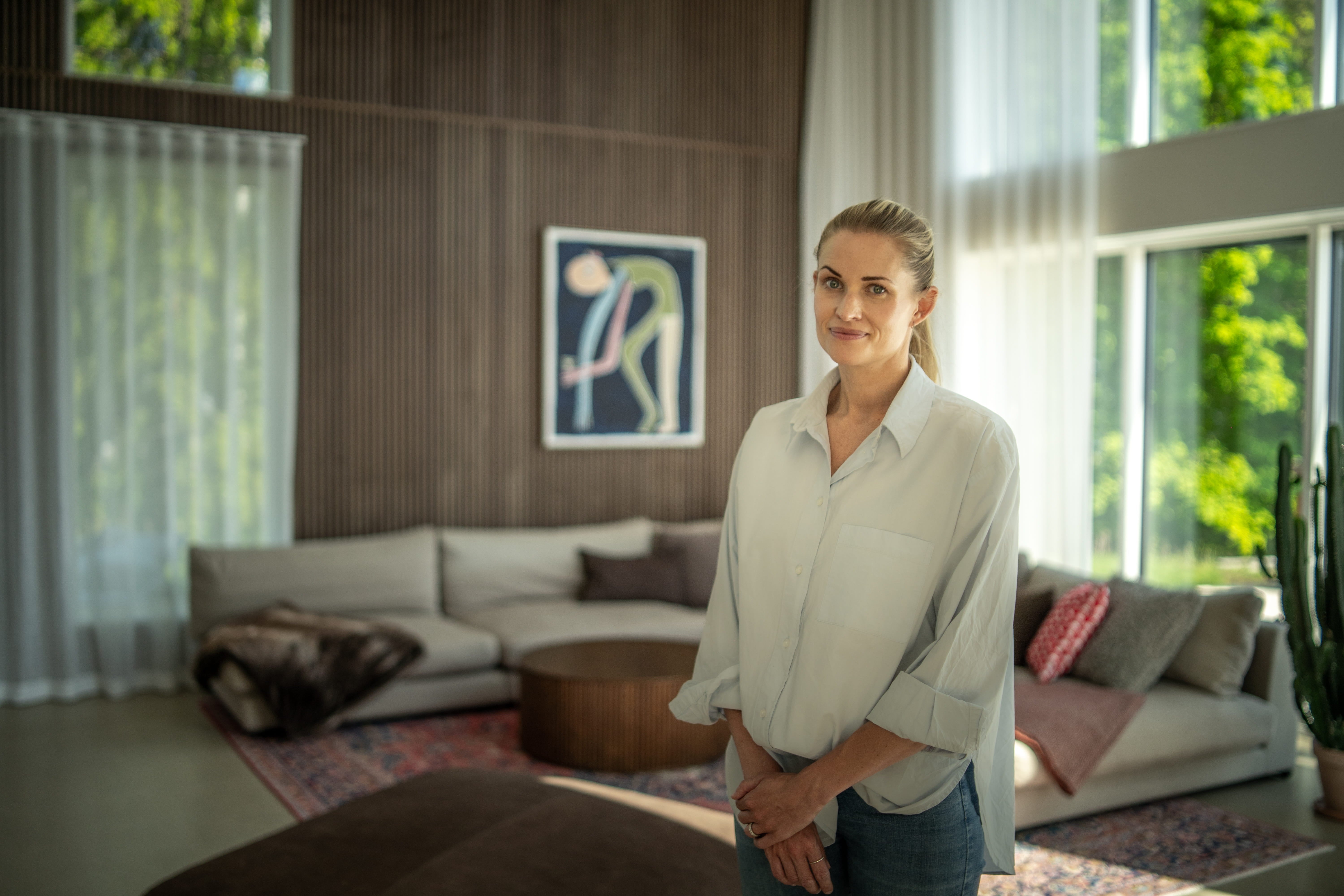 Une femme se tient dans un salon ensoleillé, avec de grandes fenêtres, des panneaux de bois acoustique nordique au mur, des œuvres dart contemporaines et des meubles minimalistes aux tons neutres.